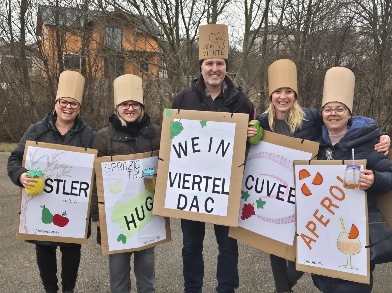 Fünf Personen stehen zusammen und tragen kegelförmige Papiermützen. Sie halten Schilder mit den Aufschriften 'We in Viertel DAC', 'Spritzig fru Hug', 'Stiler' und 'Cuvée Aperol'. Sie lächeln und scheinen für ein Foto zu posieren.