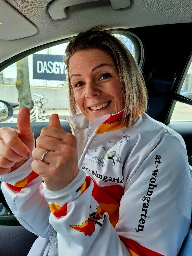 A smiling woman in a white jacket with red and yellow accents gives a thumbs up in a car. Behind her, a bicycle is parked, and a signboard with the text 'DASGY' is visible.