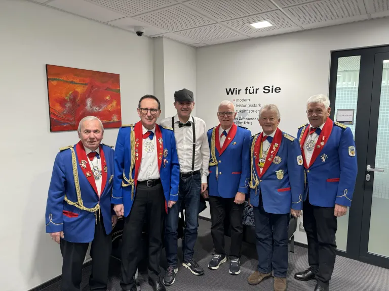 Eine Gruppe von Männern in blauen Uniformen und Hüten steht zusammen in einem Raum, ein Mann in einem weißen Hemd steht in der Mitte. Ein Gemälde befindet sich an der Wand hinter ihnen.