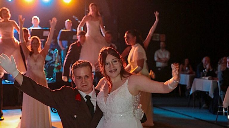 A couple dancing in a wedding reception with a band in the background, some people raising their hands and smiling.