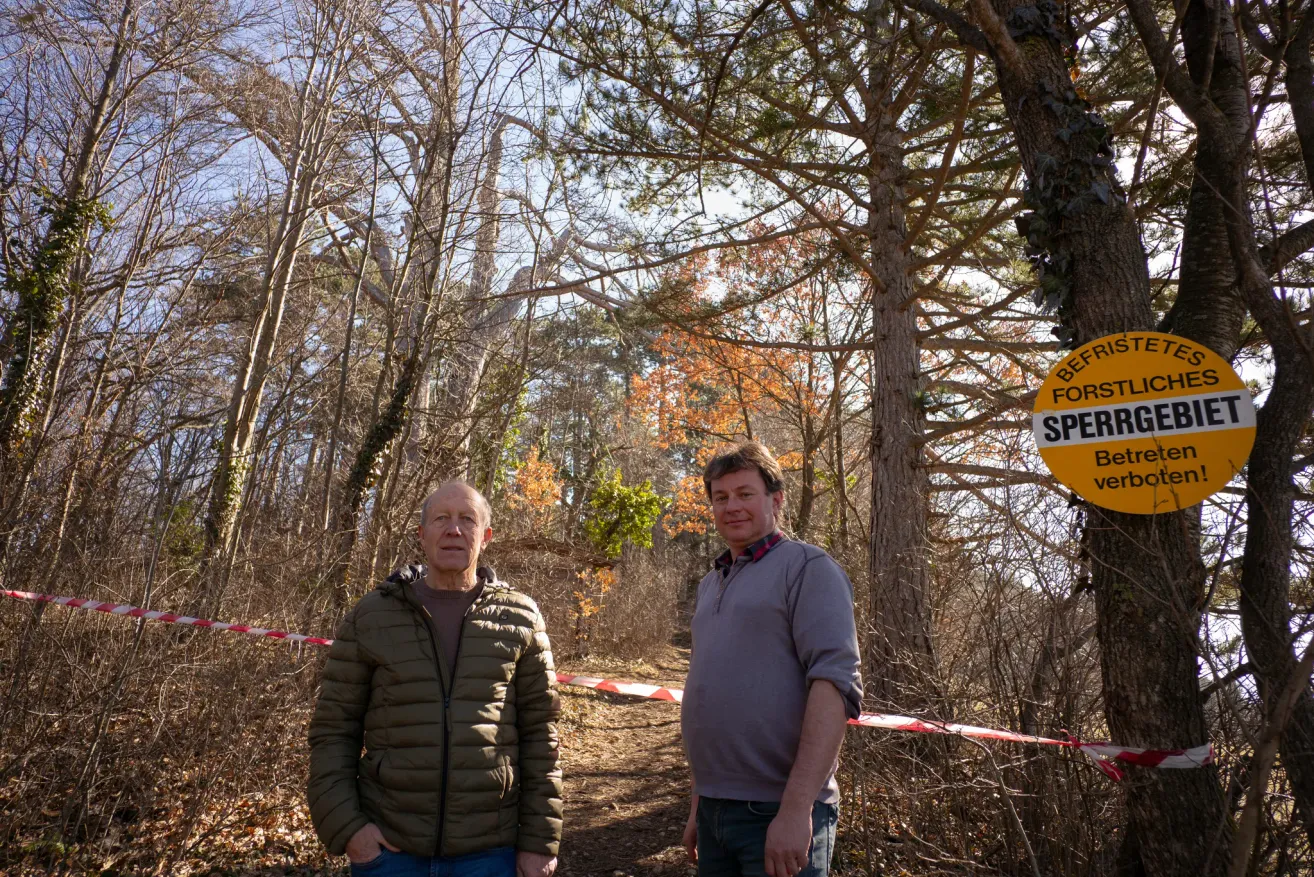 Zwei Männer stehen in einem Wald und posieren für ein Foto. Hinter ihnen befindet sich eine Barriere mit roten und weißen Streifen. Bäume mit kahlen Ästen und einigen grünen Blättern umgeben sie.