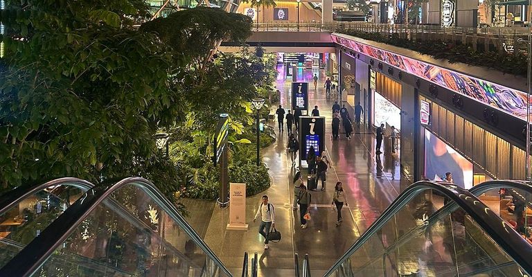 A busy indoor mall with people walking and shops along the side, illuminated with pink lights. An escalator runs on the left side.
