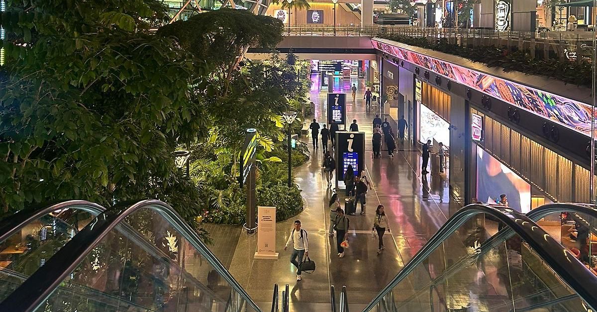 A busy indoor mall with people walking and shops along the side, illuminated with pink lights. An escalator runs on the left side.