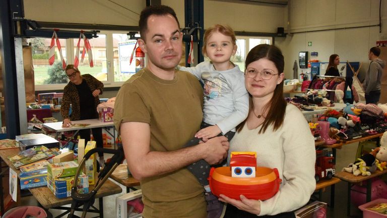 Ein Mann, eine Frau und ein kleines Mädchen stehen lächelnd zusammen im Innenbereich. Der Mann hält das Mädchen, und die Frau hält ein Spielzeugboot. Hinter ihnen steht ein Tisch mit Spielzeug und einem Feuerlöscher.