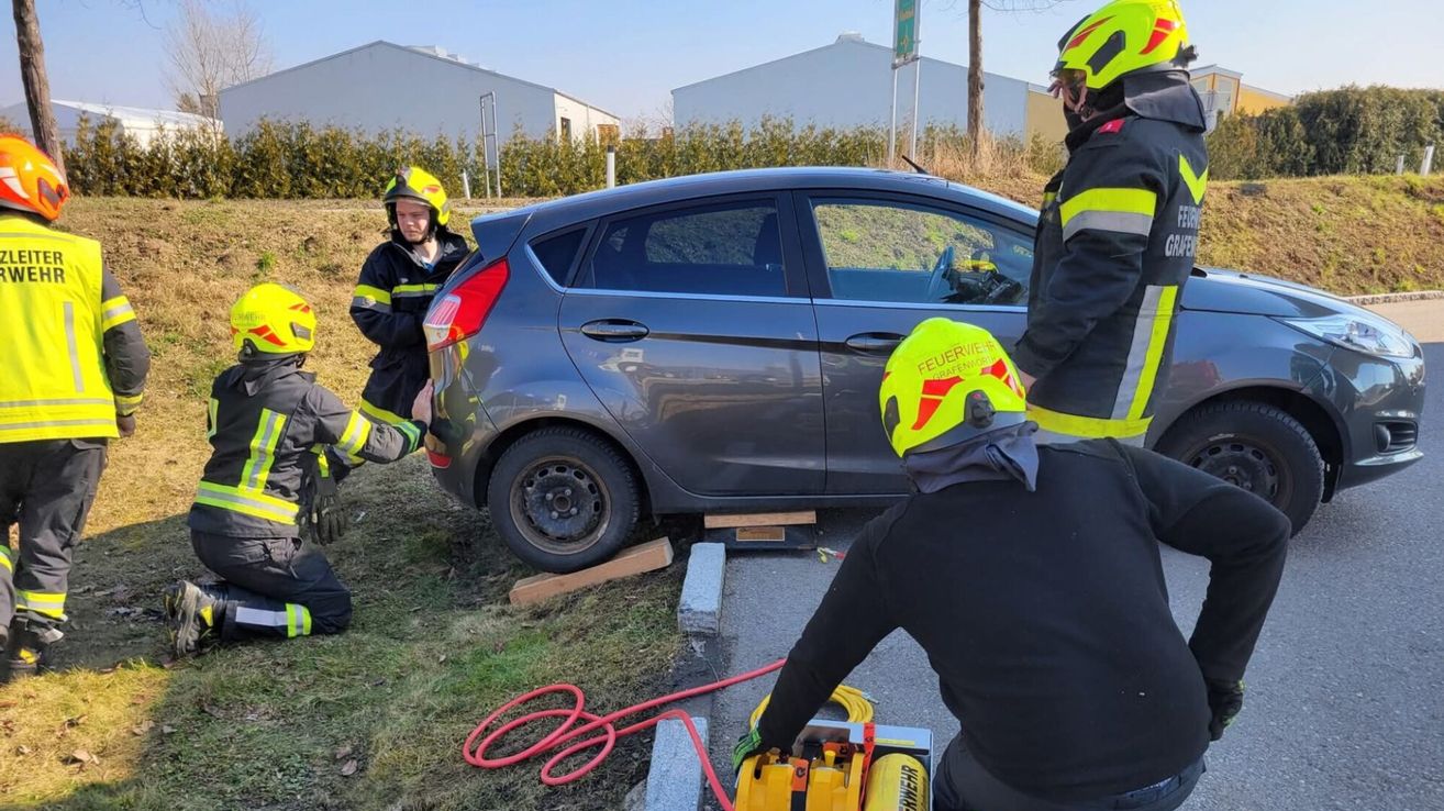A group of firefighters are working on a car, possibly extracting someone from the vehicle. One is using a hydraulic tool.
