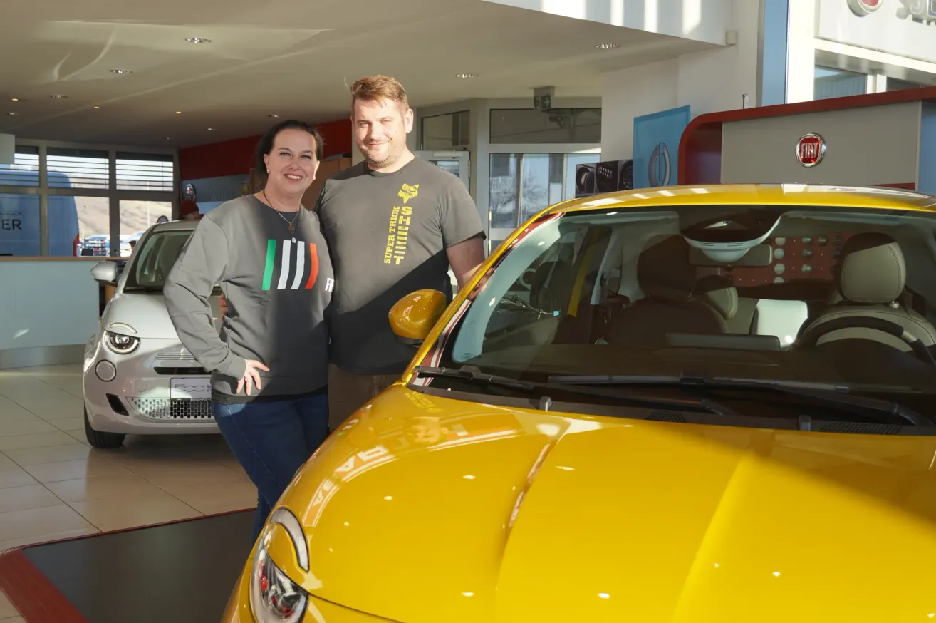 Ein Mann und eine Frau stehen neben einem gelben Auto in einem Autohaus. Die Frau trägt ein schwarzes Sweatshirt mit italienischem Flaggendesign.