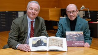 Zwei Männer sitzen an einem Holztisch mit einem offenen Buch. Der Mann auf der linken Seite trägt eine rote Krawatte, während der Mann auf der rechten Seite eine Brille trägt.