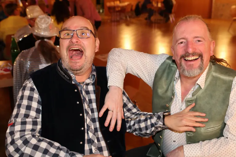 Zwei Männer lachen in einer Innenumgebung; einer in einem karierten Hemd und Weste, der andere in einem weißen Hemd mit einer grünen Weste. Sie legen die Hände auf die Schultern des anderen. Unscharfe Menschen und Tische sind im Hintergrund.