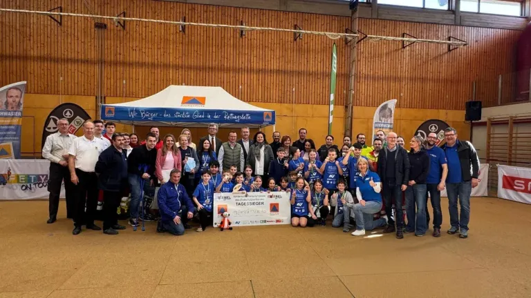 Eine Gruppe von Menschen, darunter Kinder in blauen Trikots, posiert für ein Foto unter einem Zelt mit einem Banner. Dahinter stehen Erwachsene in verschiedenen Kleidungsstücken in der Nähe von Holzwänden.