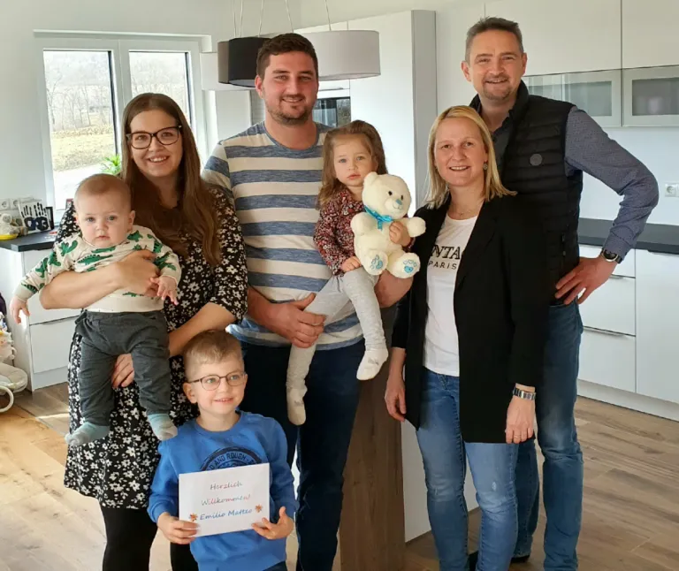 Eine sechsköpfige Familie steht in einem Wohnzimmer, ein junger Junge hält ein Schild, während ein Mädchen einen Teddybär hält. Die Erwachsenen tragen lässige Kleidung, und sie alle lächeln für die Kamera. Im Hintergrund befindet sich ein Fenster mit Blick auf ein Grasfeld.