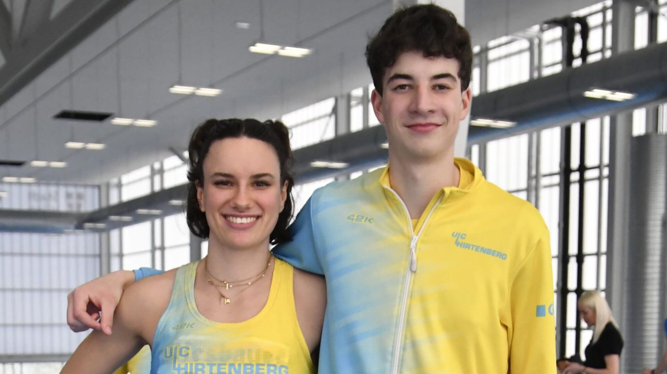 Eine lächelnde Frau und ein Mann stehen nah beieinander, beide tragen gelbe Sporttrikots mit blauen Akzenten. Sie posieren wahrscheinlich für ein Foto in einer überdachten Sportanlage.