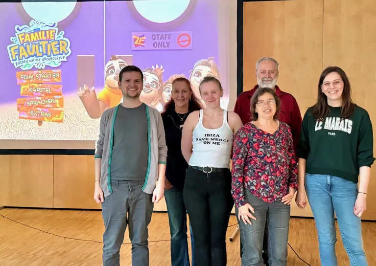 Five people stand in front of a projection screen. A sign reads 'STAFF ONLY'. One woman wears a tank top saying 'IBIZA HAVE MERCY ON ME'. The others wear casual clothes. The projection shows an animated character.