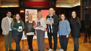 Fünf Personen stehen vor einem Banner, das 'BLICH' aufgedruckt hat. Sie halten Bücher in den Händen, wahrscheinlich für ein Foto. Eine Frau mit Brille steht rechts, während eine andere ein Buch mit 'SIRNA ADRIAN' auf dem Cover hält. Ein Mann mit langen Haaren lächelt in die Kamera.