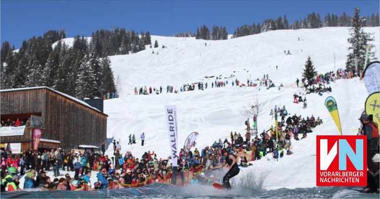 Ein Snowboarder führt einen Sprung auf einer Wasserrampe in einem Skigebiet aus, umgeben von einer großen Menge. Die verschneite Landschaft und Bäume sind im Hintergrund zu sehen.
