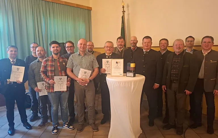 Eine Gruppe von Männern steht um einen Tisch mit Auszeichnungen und Zertifikaten. Einige halten Zertifikate, während andere Auszeichnungen halten. Hinter ihnen befindet sich eine Flagge und ein grüner Vorhang.