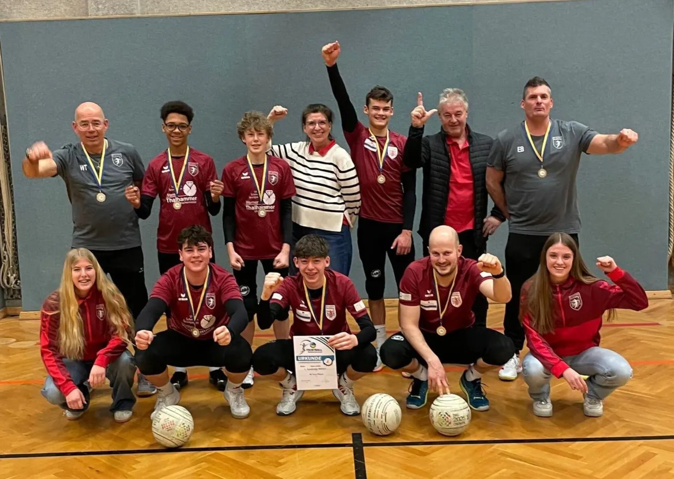 Eine Gruppe junger Athleten, die rote Trikots und Medaillen tragen, posiert für ein Foto auf einem Holzboden. Einige hocken, während andere hinter ihnen stehen und alle lächeln. In der Mitte wird ein Zertifikat mit 'URKUNDEN' gehalten.