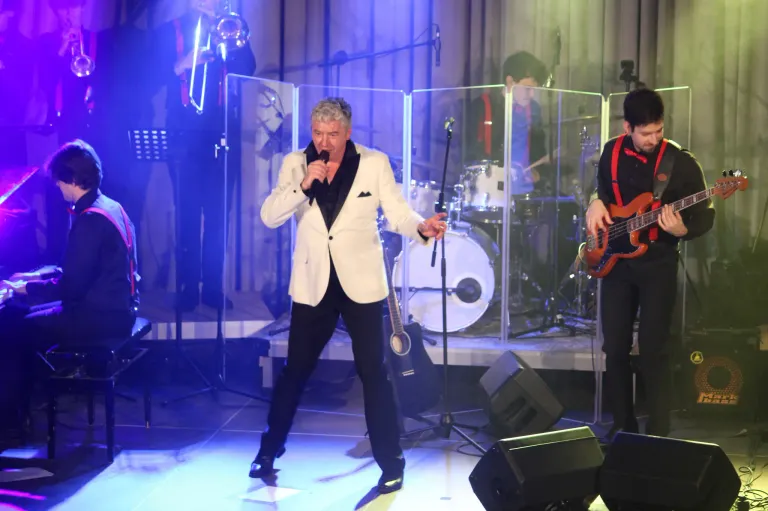 Ein Mann tritt auf der Bühne auf, singt in ein Mikrofon und trägt einen weißen Anzug. Hinter ihm spielt ein Schlagzeuger, und ein Gitarrist steht rechts. Die Bühne ist mit blauem Licht beleuchtet.