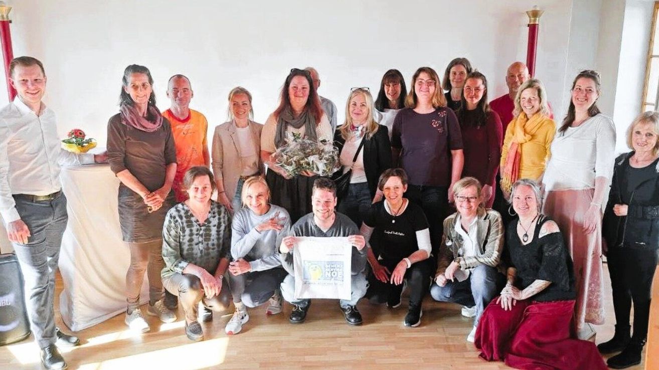 A group of adults posing for a photo in a room, some are kneeling, and one man holds a white banner with text. A woman holds a bouquet. They are all smiling.