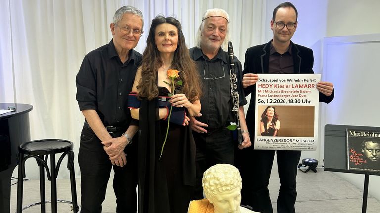 Vier Personen stehen neben einer Statue und lächeln. Die Frau hält eine Rose, während der Mann auf der rechten Seite ein Schild hält. Sie tragen dunkle Kleidung. Der Mann in der Mitte hält eine Klarinette. Der Mann auf der linken Seite trägt eine Brille.