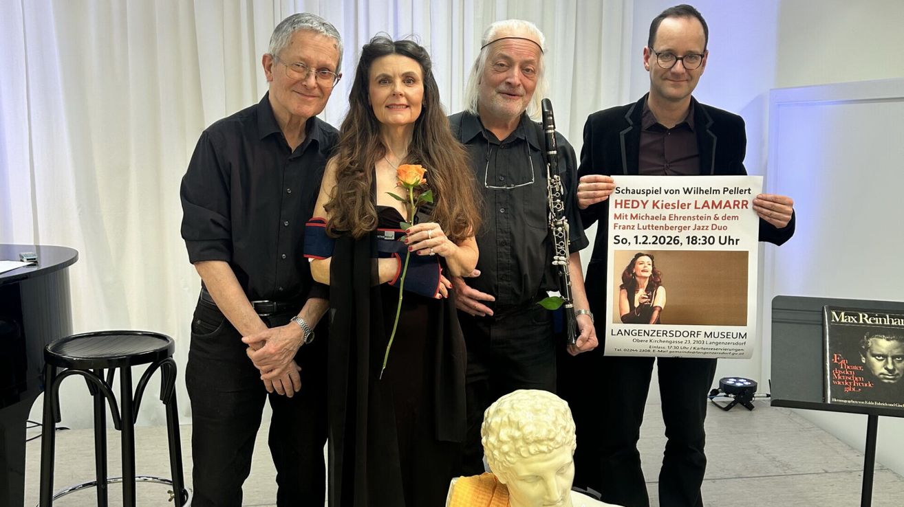 Vier Personen stehen neben einer Statue und lächeln. Die Frau hält eine Rose, während der Mann auf der rechten Seite ein Schild hält. Sie tragen dunkle Kleidung. Der Mann in der Mitte hält eine Klarinette. Der Mann auf der linken Seite trägt eine Brille.