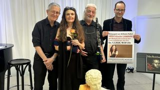 Vier Personen stehen neben einer Statue und lächeln. Die Frau hält eine Rose, während der Mann auf der rechten Seite ein Schild hält. Sie tragen dunkle Kleidung. Der Mann in der Mitte hält eine Klarinette. Der Mann auf der linken Seite trägt eine Brille.