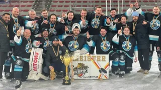 Eine Eishockeymannschaft in schwarzen und weißen Trikots posiert für ein Foto auf einer Eisbahn. Sie halten einen Pokal und Medaillen.