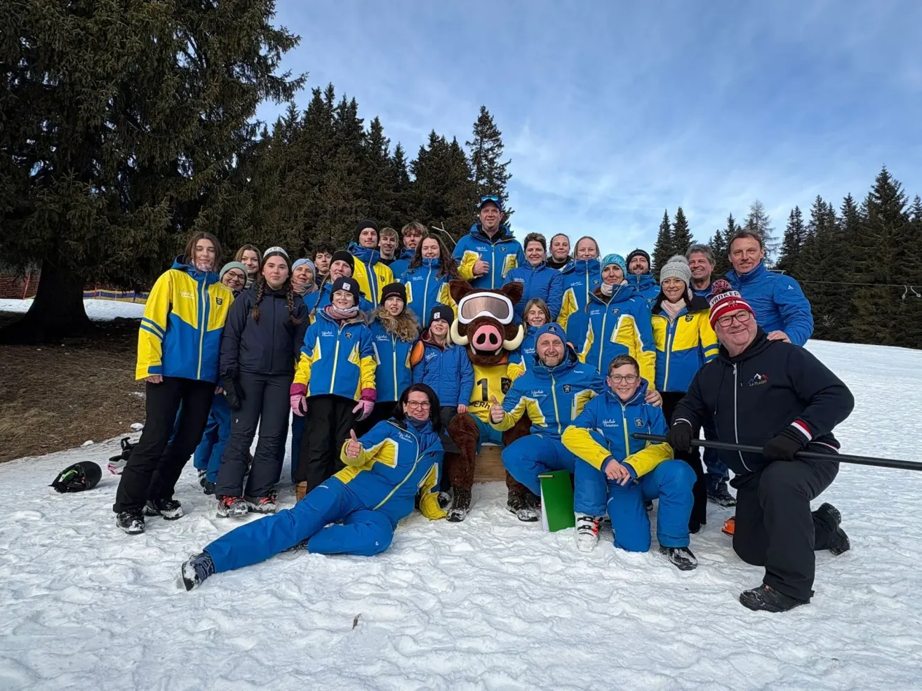 Eine Gruppe von Menschen, die in blauen und gelben Skianzügen und -jacken gekleidet sind, posiert für ein Foto im Schnee mit einem Maskottchen in der Mitte. Hinter ihnen stehen Bäume im Hintergrund.