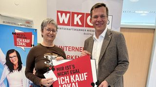 Eine Frau und ein Mann stehen vor einem Banner. Die Frau hält ein Zertifikat und eine Karte. Der Mann hält ein rotes Schild mit weißem Text. Das Banner hat ein Logo, das 'WKC Wirtschaftskammer Nieder' liest.