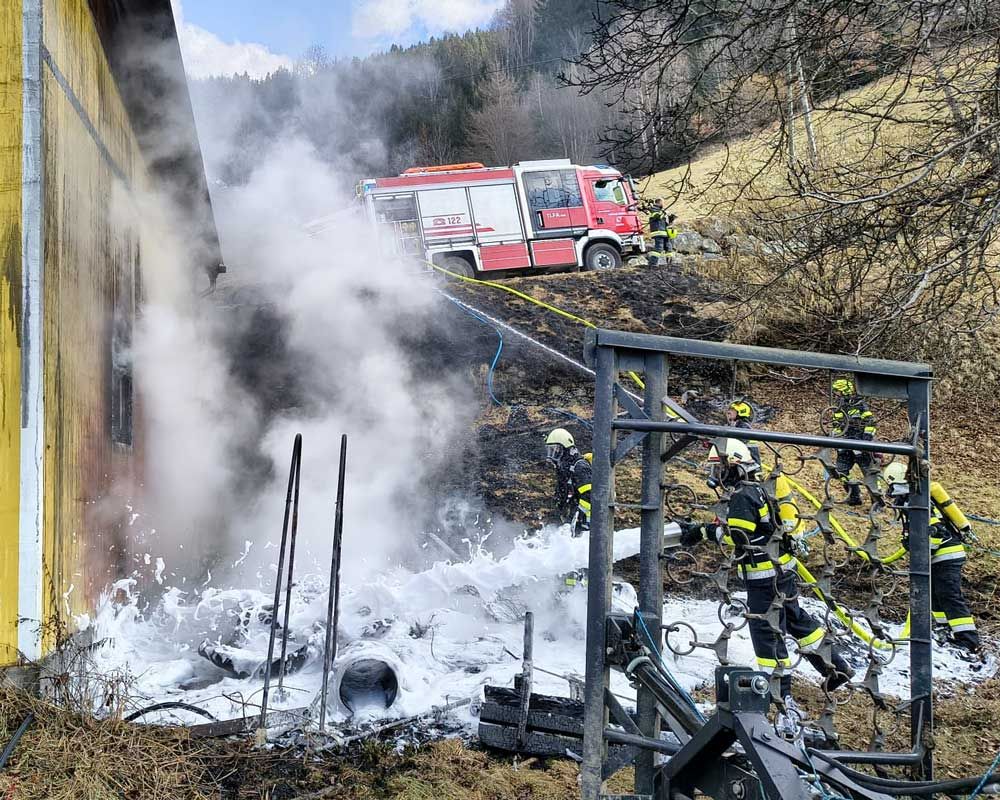 Feuerwehrleute sind bei einem Gebäudebrand im Einsatz und verwenden einen Schlauch, um Wasser auf die Flammen zu sprühen. Rauch steigt aus dem Gebäude auf, und mehrere Feuerwehrleute sind mit Schutzausrüstung ausgestattet.