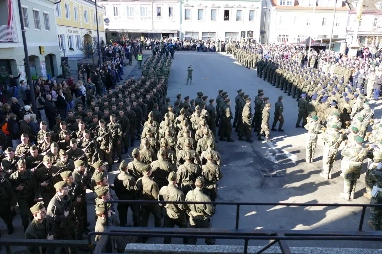 A large military parade takes place in an urban setting, with soldiers in green uniforms lined up on both sides of the street. Spectators watch from the sidewalks and buildings line the street.