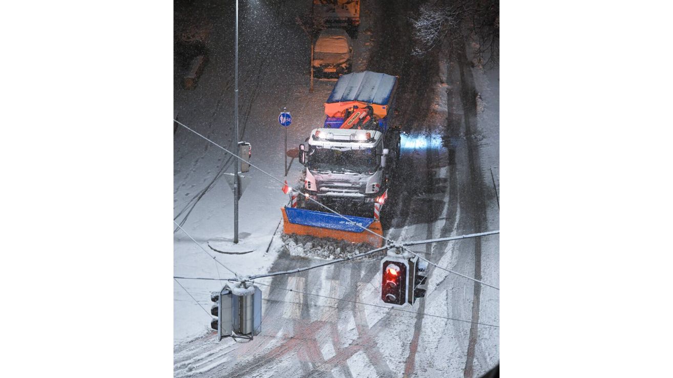 Ein Schneepflug räumt den Schnee von der Straße. Er pflügt durch den Schnee, wobei eine Ampel und ein Verkehrsschild sichtbar sind.