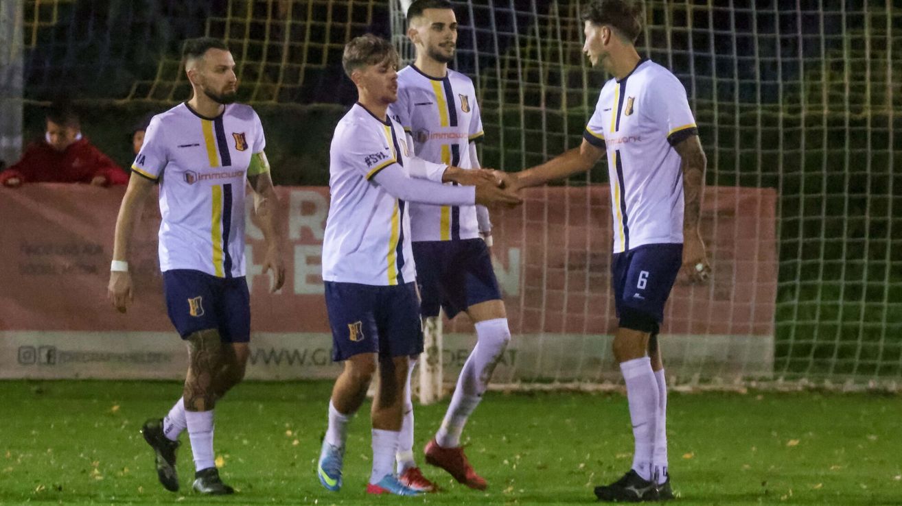 Soccer players in white uniforms celebrate a goal on a grassy field with a goalpost in the background.