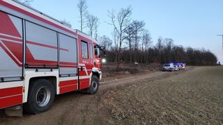 Ein Feuerwehrauto steht auf einem Feldweg in einem Waldgebiet. Das Auto ist rot mit weißen Streifen. Der Weg ist von kahlen Bäumen umgeben. In der Nähe sind zwei Polizeiautos geparkt.
