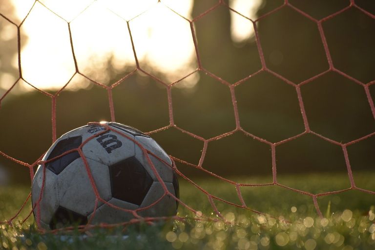 A soccer ball is in the goal net, caught between the red strings, under a bright sun.