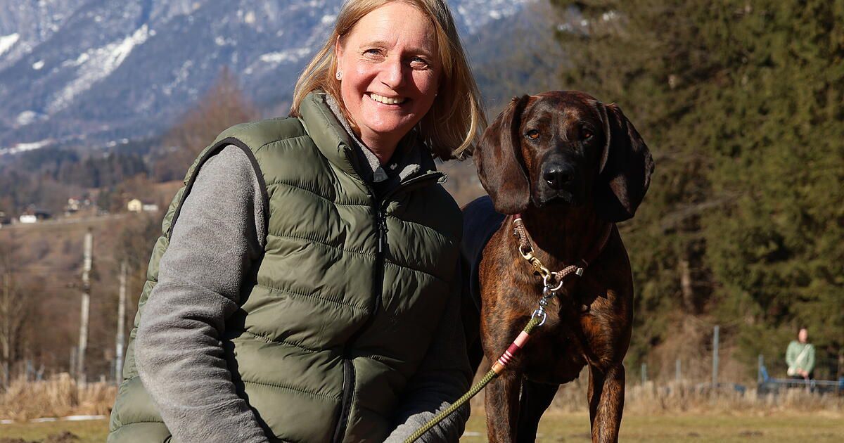 A woman in a green vest stands with a brown dog, smiling and looking at the camera, with snowy mountains in the background.