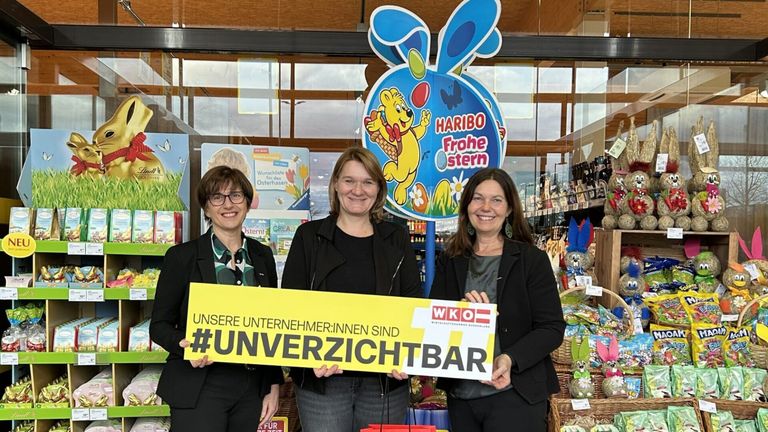 Drei Frauen stehen vor einem Geschäft und halten ein gelbes Schild. Auf dem Schild steht 'Unsere Unternehmen:innen sind #unverzichtbar.' Dahinter befindet sich ein großes blaues Schild mit einem gezeichneten Bären, der einen Korb mit Eiern hält und die Worte 'HARIBO Frohe stern' darauf stehen.