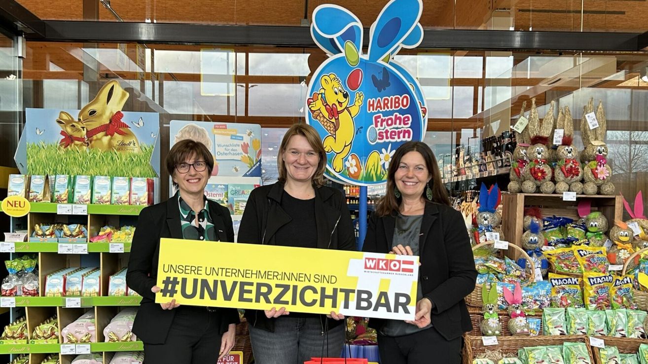 Drei Frauen stehen vor einem Geschäft und halten ein gelbes Schild. Auf dem Schild steht 'Unsere Unternehmen:innen sind #unverzichtbar.' Dahinter befindet sich ein großes blaues Schild mit einem gezeichneten Bären, der einen Korb mit Eiern hält und die Worte 'HARIBO Frohe stern' darauf stehen.