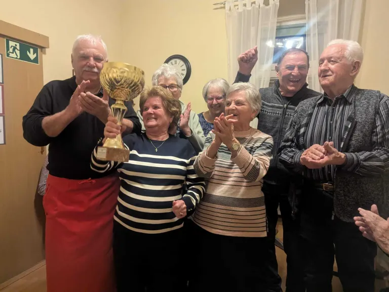 Eine Gruppe älterer Menschen feiert mit einem Pokal, einige klatschen, in einem Raum mit Uhr und Vorhängen.