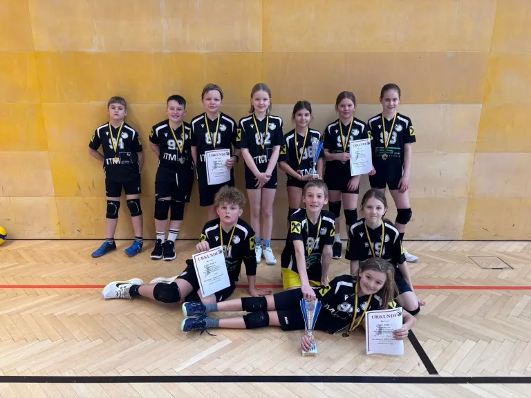 Eine Gruppe junger Athleten posiert für ein Foto mit Medaillen und Urkunden. Sie tragen Sportuniformen mit Nummern und stehen in einer Reihe, zwei knien im Vordergrund. Sie befinden sich in einer Sporthalle.