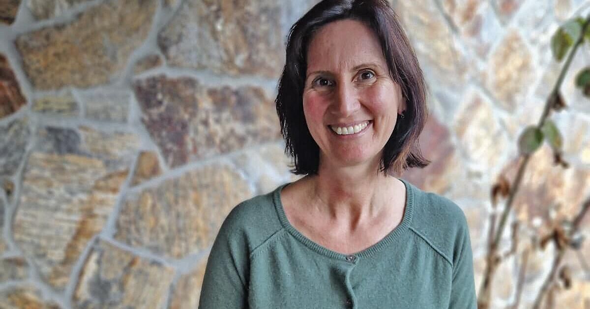 A woman with short brown hair smiles brightly. She wears a green shirt and stands against a stone wall.