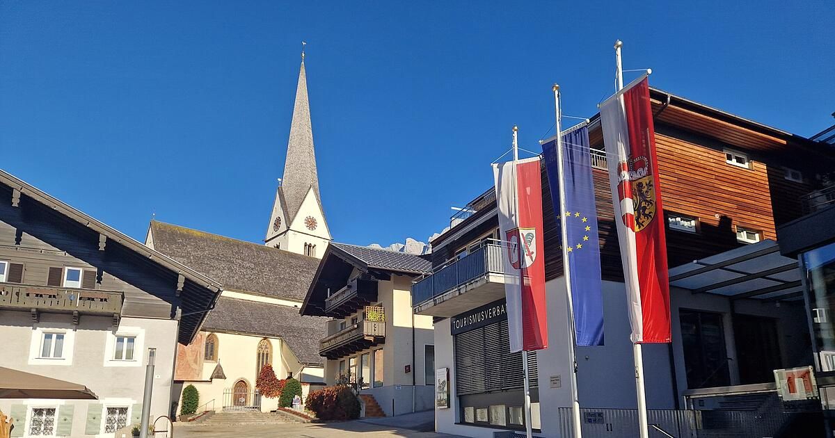 A picturesque village scene with a church featuring a steeple and clock tower. Several flags are displayed outside a building with the text 'TOURISMUSVERBA'. The background is a clear blue sky.