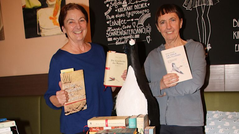 Two women stand behind a table, smiling and holding books. One woman holds a penguin statue. The books they hold have German text on them.