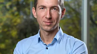 Ein Mann in einem blauen Hemd mit ernster Miene steht mit verschränkten Armen vor einem Fenster mit Bäumen draußen.