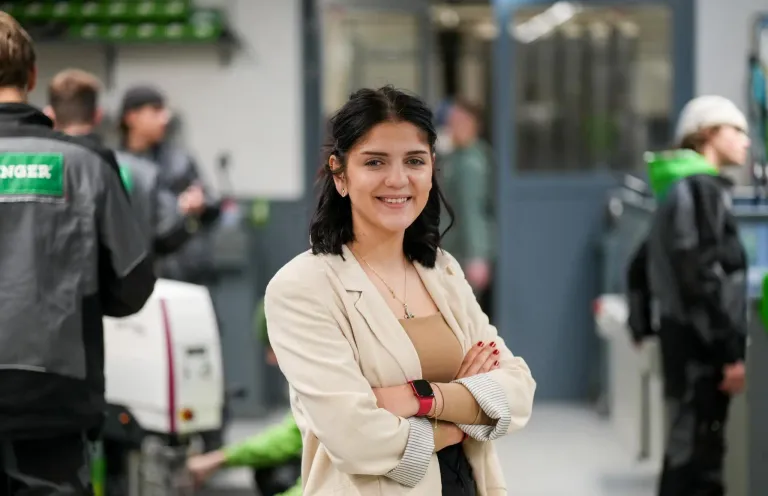 Eine Frau mit dunklen Haaren lächelt und posiert für ein Foto in einer Werkstatt. Sie trägt einen hellen Blazer und eine Halskette. Hinter ihr sind verschwommene Figuren in einem Raum mit weißen Wänden.