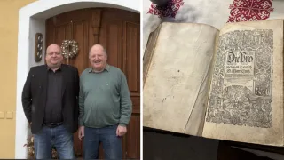 Zwei Männer stehen vor einer Holztür mit einem Kranz, einer raucht. Daneben liegt ein altes, beschädigtes Buch auf einem weißen Tuch mit roten Mustern.