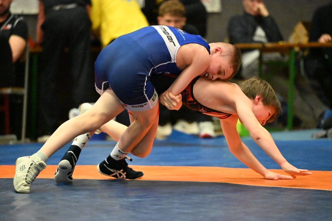 Two young wrestlers compete in a match on a blue mat. One wears a blue uniform, the other a red and black one. Spectators watch from the background.