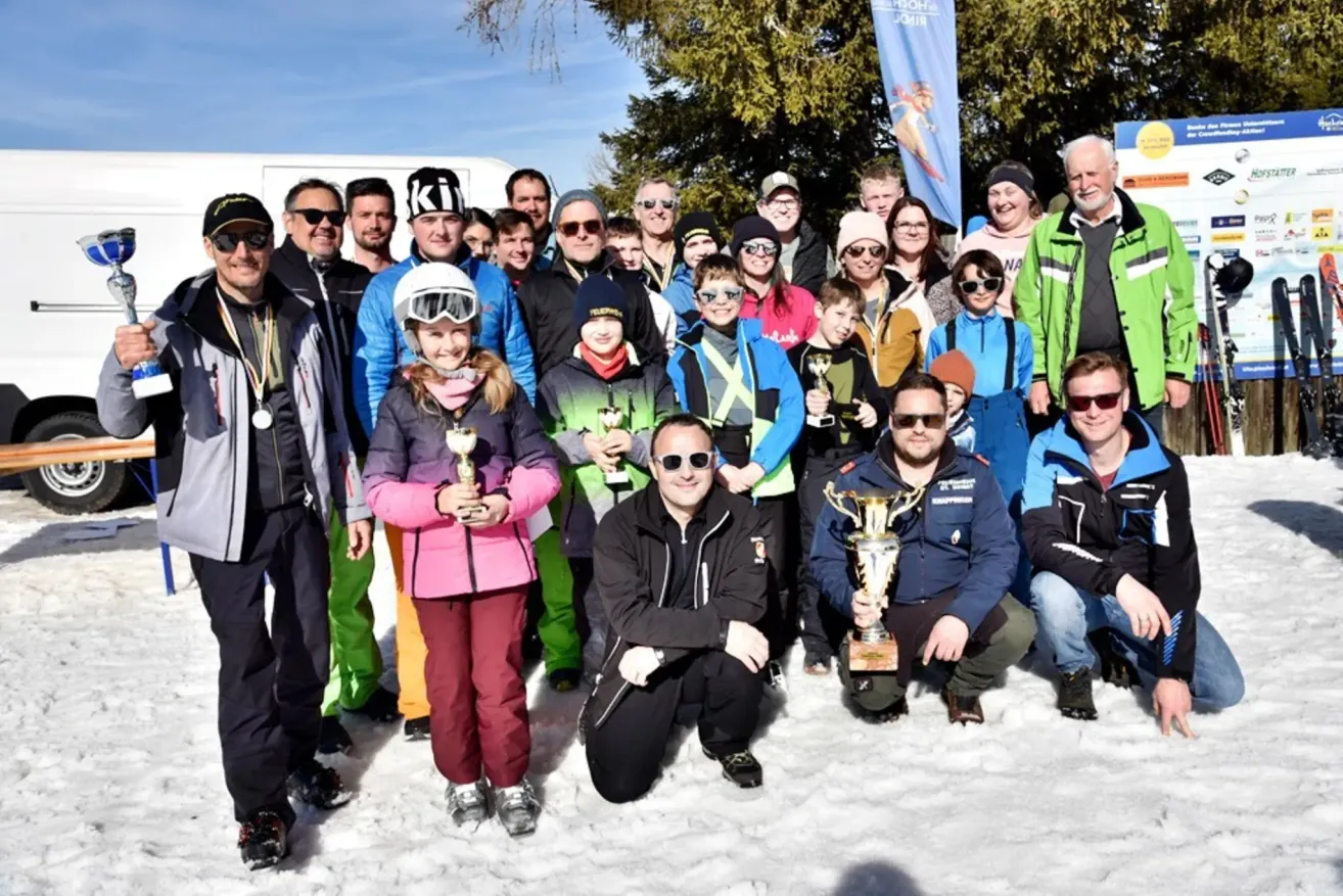 Eine Gruppe von Menschen, Erwachsene und Kinder, posiert für ein Foto mit Trophäen. Sie tragen Winterkleidung und Sonnenbrillen. Sie stehen und knien im Schnee, mit Hintergrund von Bäumen und einem Banner.