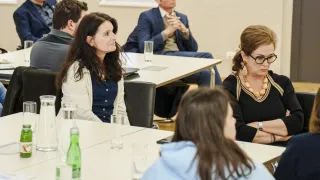 Eine Gruppe von Menschen sitzt in einem Konferenzraum um einen Tisch herum. Flaschen, Gläser und Teller sind auf dem Tisch. Der Mann rechts trägt eine Brille und hält seine Hände zusammen. Die Frau in der Mitte schaut nach links.
