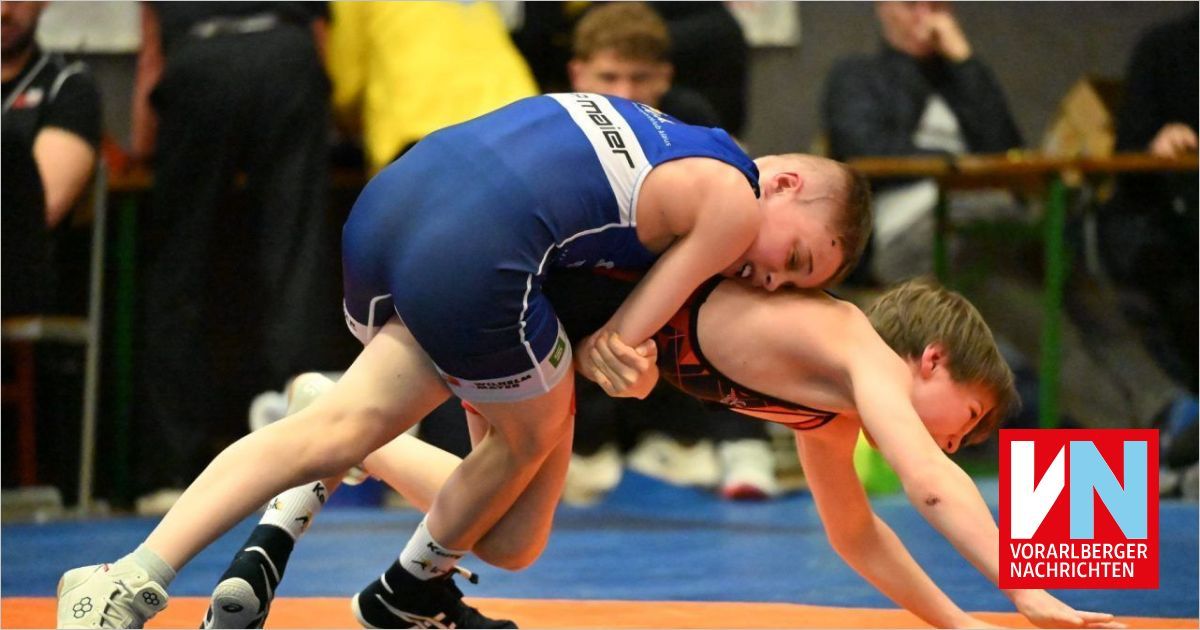 Two young male wrestlers compete in a blue-ringed arena. One in blue holds the other in red, both are in mid-action, showcasing intense focus. Spectators watch in the background.