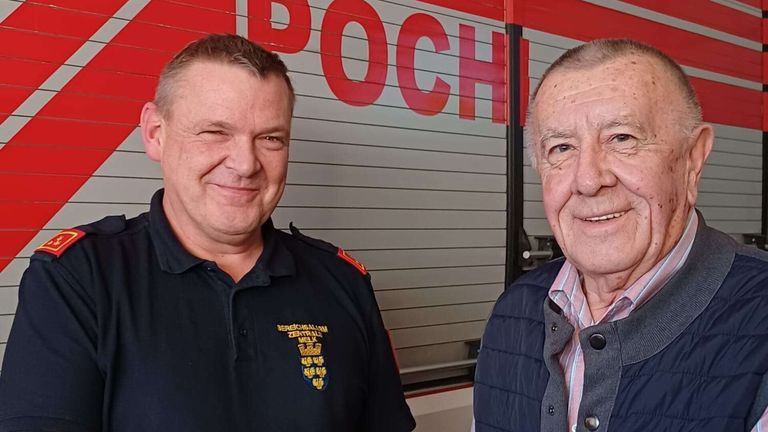 Zwei Männer lächeln und posieren für ein Foto vor einem Feuerwehrauto. Ein Mann trägt eine schwarze Uniform, der andere eine blaue Weste.